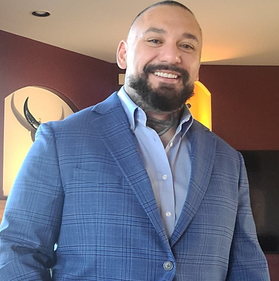 A smiling man with a beard and tattoos on his neck wears a light blue plaid suit jacket over a blue dress shirt, standing indoors with warm lighting and artwork in the background.